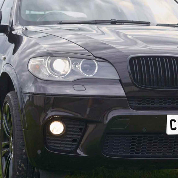 A dark SUV with glowing headlights is parked on grass, featuring reflective paint and a front license plate reading "CARBON." The sky is cloudy.