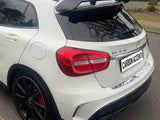 A white sporty car, possibly a Mercedes-Benz GLA 45 with tinted windows and a roof spoiler, is parked on a suburban street. The license plate reads 