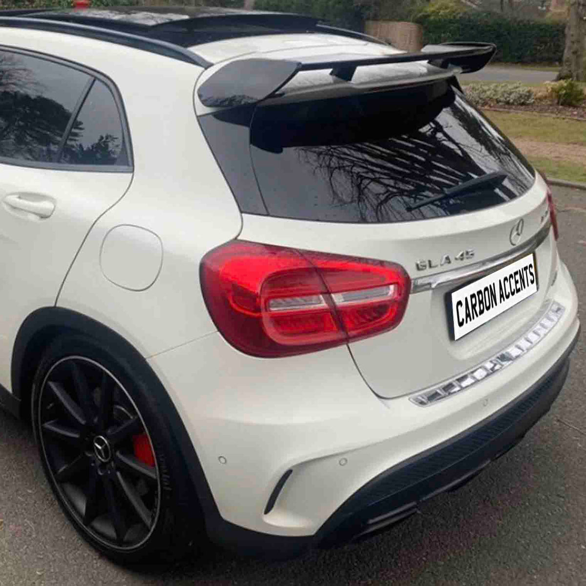 A white sporty car, possibly a Mercedes-Benz GLA 45 with tinted windows and a roof spoiler, is parked on a suburban street. The license plate reads "CARBON ACCENTS."