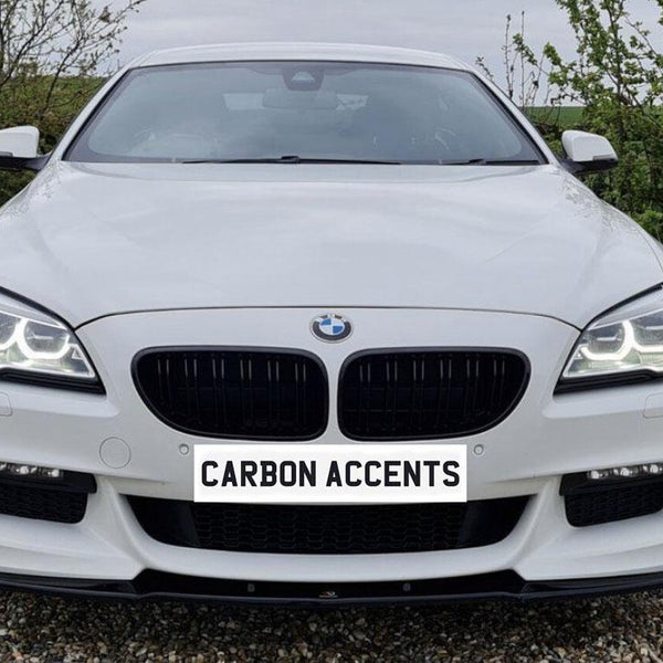 A white car with illuminated headlights is parked on gravel, surrounded by greenery. The front license plate reads "CARBON ACCENTS."