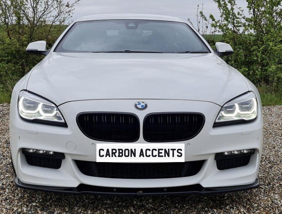 A white car with illuminated headlights is parked on gravel, surrounded by greenery. The front license plate reads "CARBON ACCENTS."