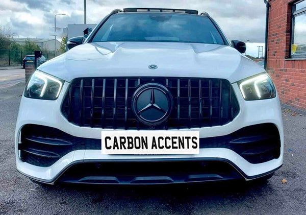 A white SUV with illuminated headlights is parked on a paved area beside a brick building. The front license plate displays "CARBON ACCENTS." The sky is overcast.