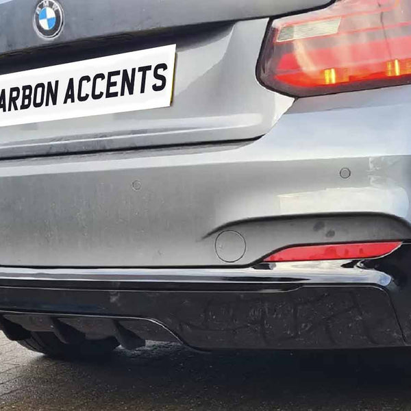 A silver car displays a "CARBON ACCENTS" license plate, featuring dual exhaust pipes and red taillights. It is parked on a textured surface, with a building in the background.