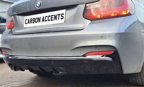 A silver car displays a "CARBON ACCENTS" license plate, featuring dual exhaust pipes and red taillights. It is parked on a textured surface, with a building in the background.