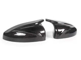 Two sleek, black carbon fiber car mirror covers rest on a plain white surface, reflecting light and showcasing their glossy, patterned finish.