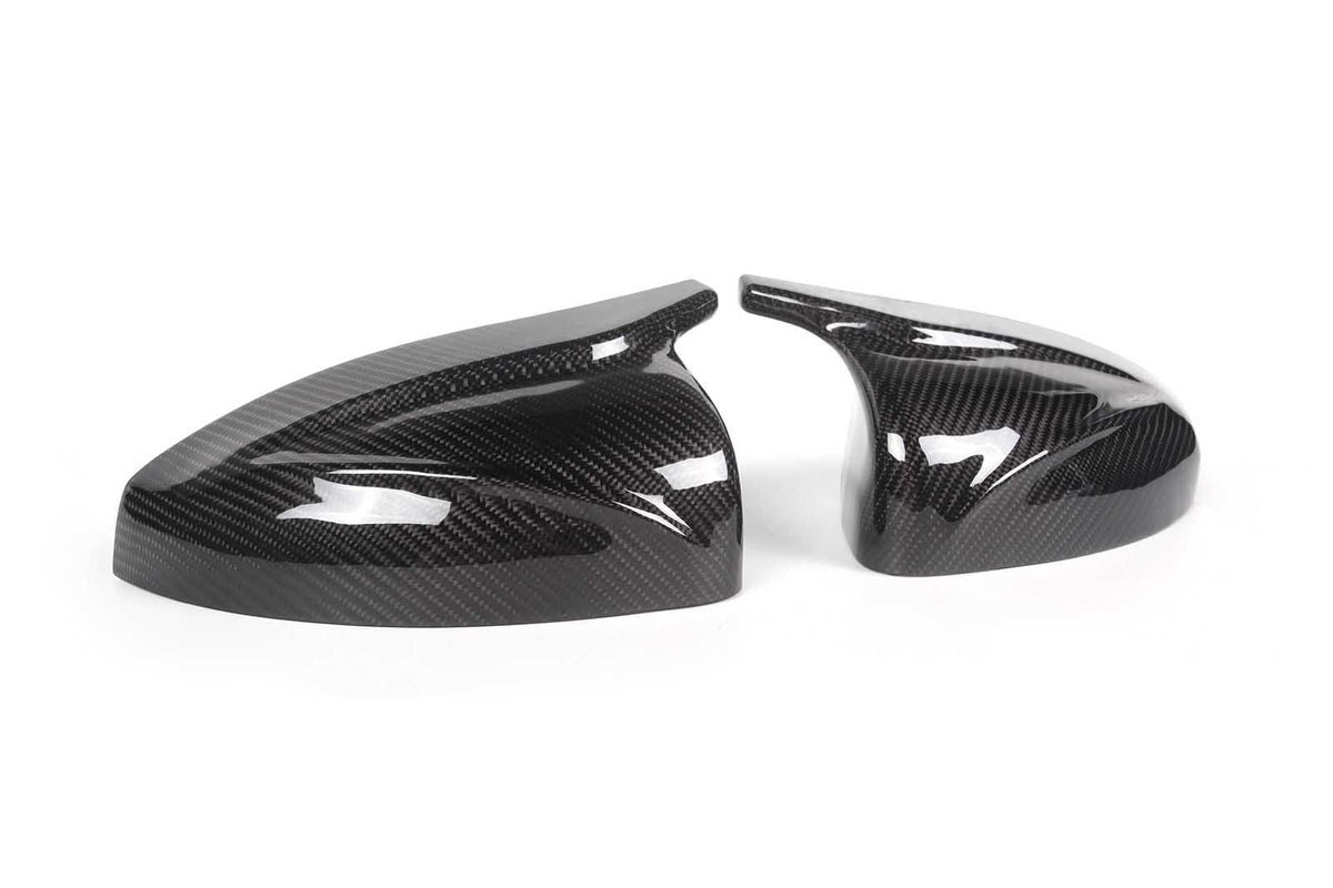 Two sleek, black carbon fiber car mirror covers rest on a plain white surface, reflecting light and showcasing their glossy, patterned finish.