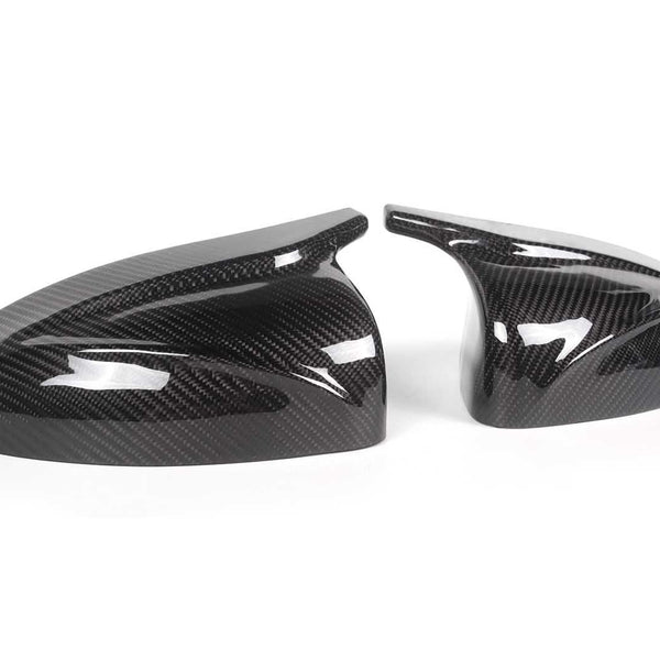 Two sleek, black carbon fiber car mirror covers rest on a plain white surface, reflecting light and showcasing their glossy, patterned finish.