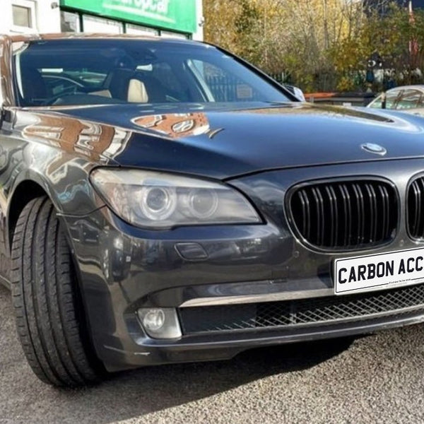 Car parked outside a building with "CARBON ACCENTS" on the license plate, surrounded by other vehicles and trees. The car has a glossy, dark finish and prominent front grille.