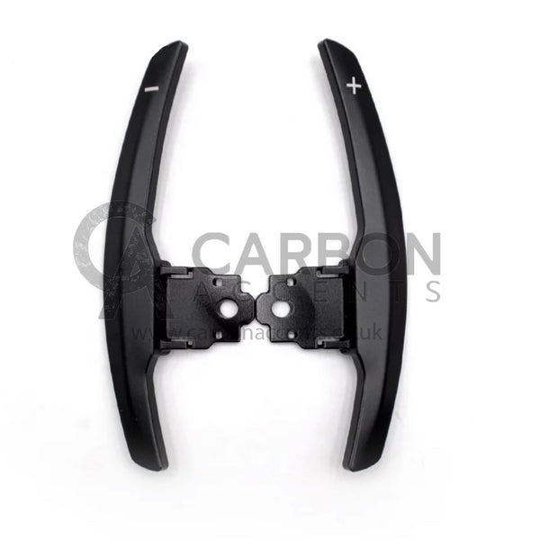 Paddle shifters with plus and minus symbols attached to a black base, used for car gear shifting. Background is plain white with faint text reading "CARBON" and "www.carbonac.uk".