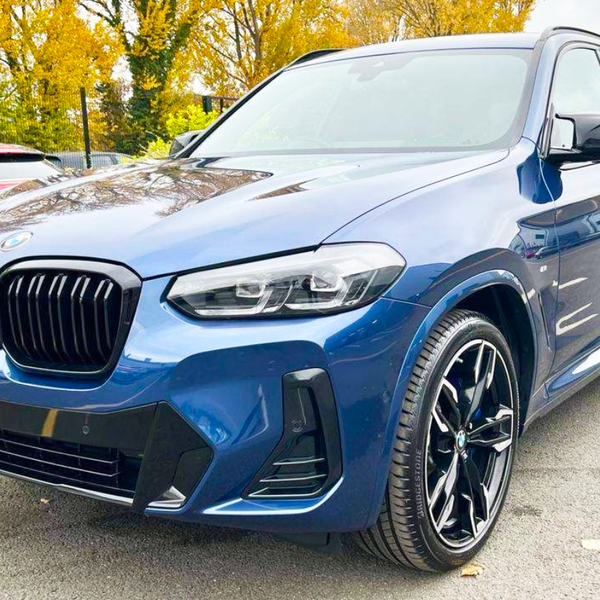 A blue SUV is parked on a paved surface, featuring a prominent black grille and sleek design. Autumn trees with golden leaves form the backdrop, alongside another parked vehicle.