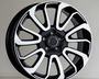 A black and white alloy wheel with ten curved spokes sits against a plain, light-gray background, resting on a flat surface.