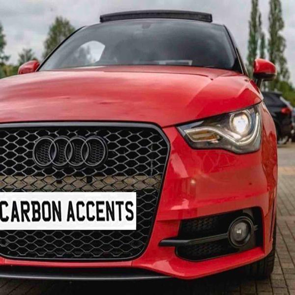A red car with an Audi logo prominently displays a license plate reading "CARBON ACCENTS," parked on a paved surface surrounded by blurred trees and another vehicle in the background.