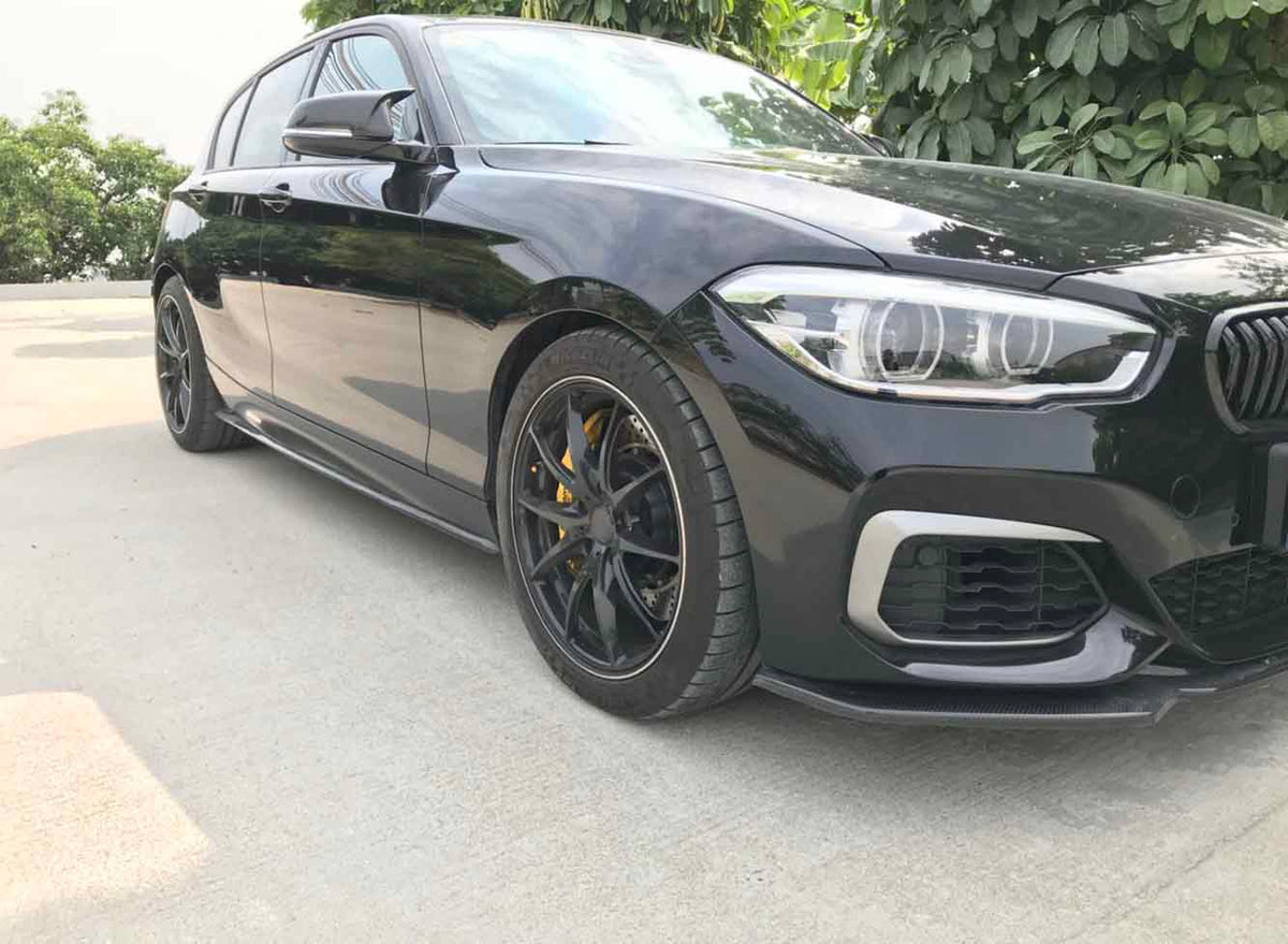 A sleek black car with sporty wheels and yellow calipers is parked on a concrete surface, surrounded by lush green foliage and trees in the background.