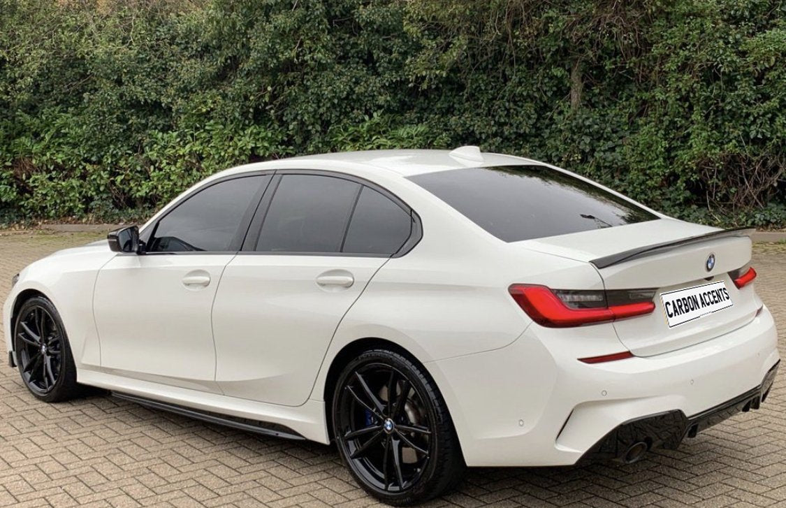 A white BMW sedan is parked on a paved surface, with tinted windows and black rims. Behind it, a dense green foliage forms the background. License plate displays "CARBON ACCENTS."