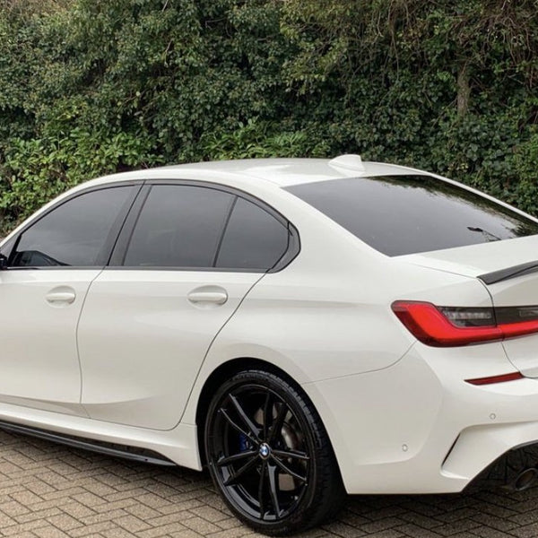 A white BMW sedan is parked on a paved surface, with tinted windows and black rims. Behind it, a dense green foliage forms the background. License plate displays "CARBON ACCENTS."