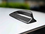 Carbon fiber shark fin antenna is mounted on a smooth, glossy white car roof, with a blurred green and gray background, suggesting an outdoor setting. No text present.