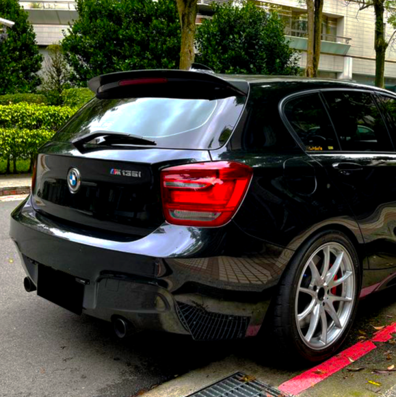 A black BMW M135i is parked on a street beside a hedge. The taillights glow, and the polished silver wheels stand out against the green foliage behind.