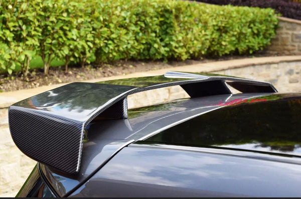 A carbon fiber car spoiler is mounted on the rear of a dark vehicle, surrounded by a neatly trimmed hedge and stone wall in a garden setting.
