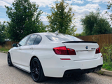 A white BMW M Sport sedan is parked on a gravel road beside a wooden fence, surrounded by trees under a partly cloudy sky.