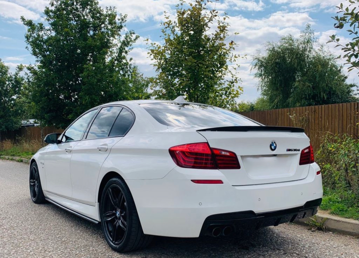 A white BMW M Sport sedan is parked on a gravel road beside a wooden fence, surrounded by trees under a partly cloudy sky.
