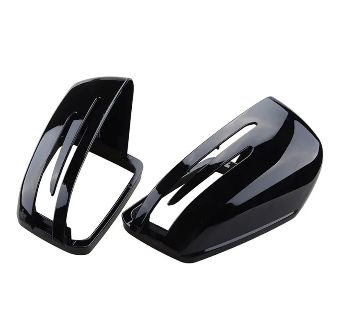 Two glossy black car mirror covers lie on a plain white surface. One is positioned upright, while the other is tilted, displaying their aerodynamic design and sleek finish.