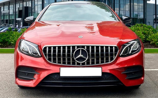 A red car with a distinctive grille and emblem is parked outdoors. Office buildings are visible in the background, along with greenery on either side.
