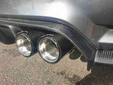 Car exhaust pipes made of shiny metal protrude from a carbon fiber rear diffuser, positioned above a gravel surface, with sunlight reflecting off the vehicle's body.