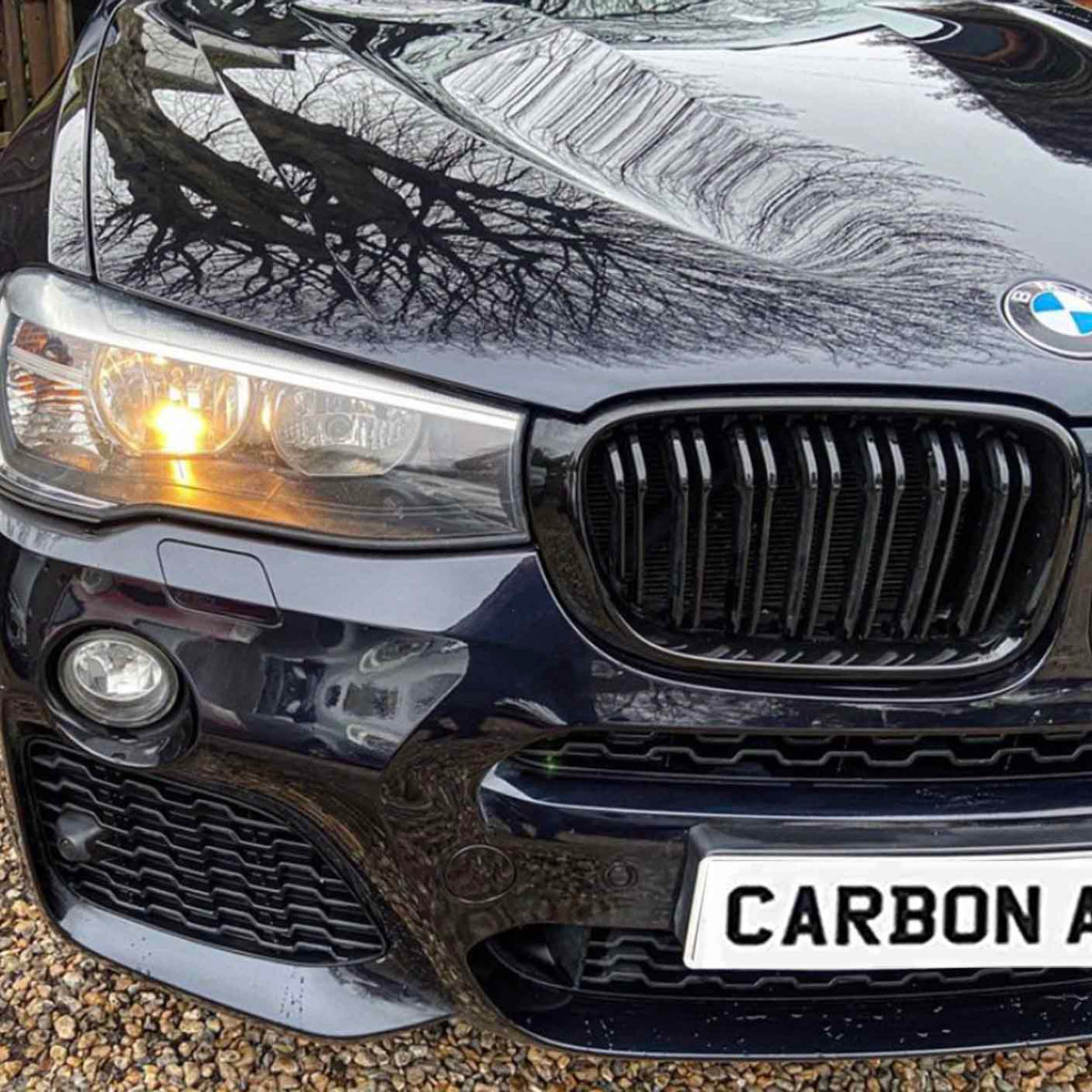 A black car's front grille and headlight are visible, with a tree's reflection on the hood. The license plate reads "CARBON AC." It is parked on gravel.