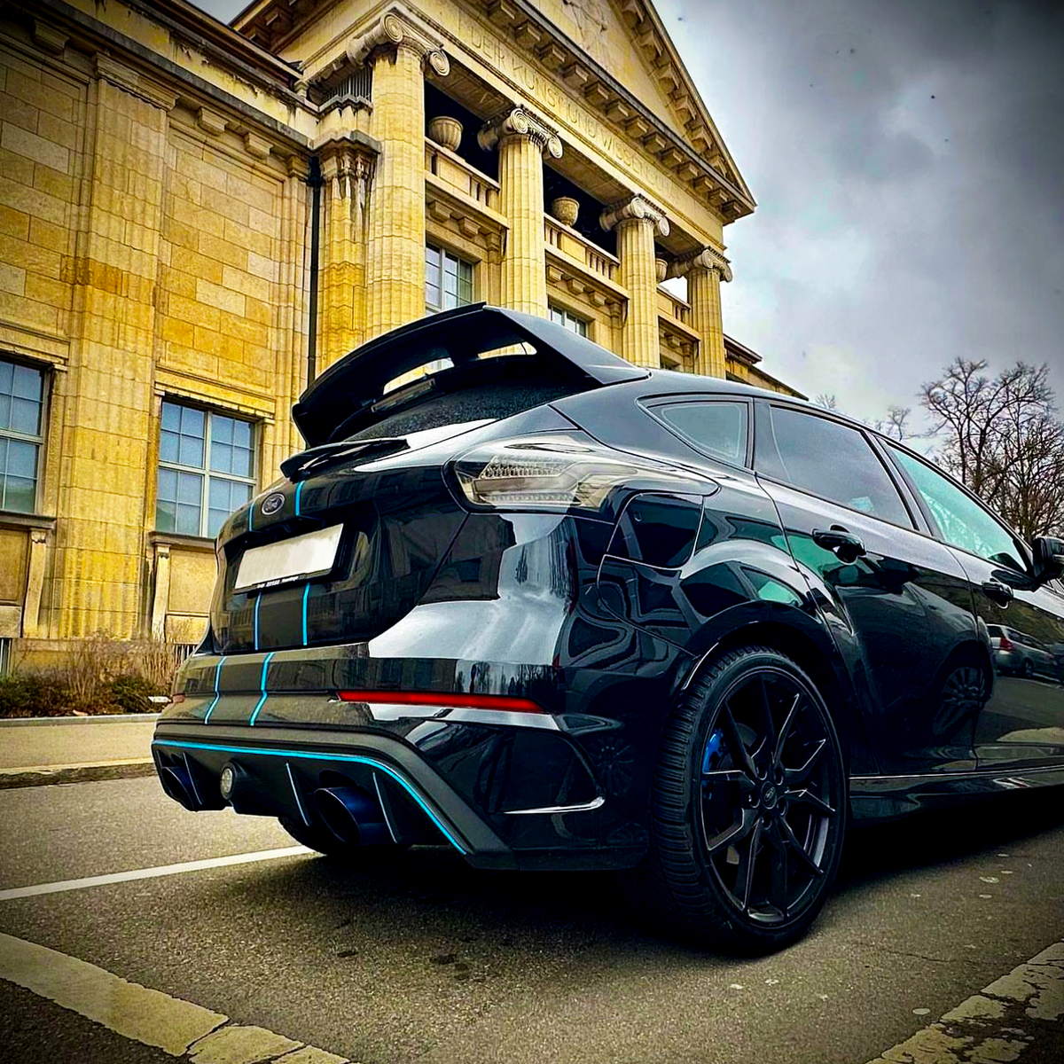 A sleek black car parked on a street in front of a neoclassical building with tall columns and a pediment. Sky is overcast, adding to the dramatic atmosphere.