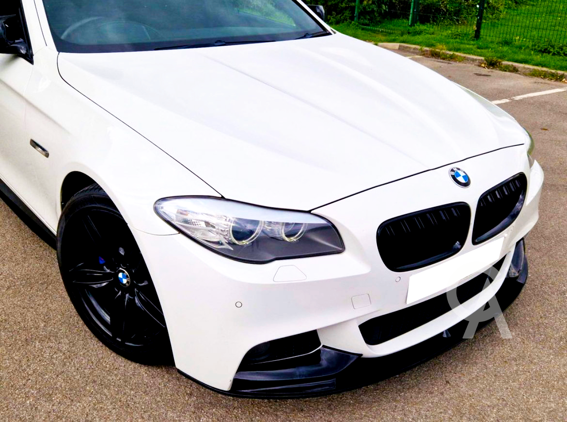 White BMW car, parked on asphalt, featuring black rims and a prominent front grille. It's situated in a parking lot bordered by green grass and a wire fence.