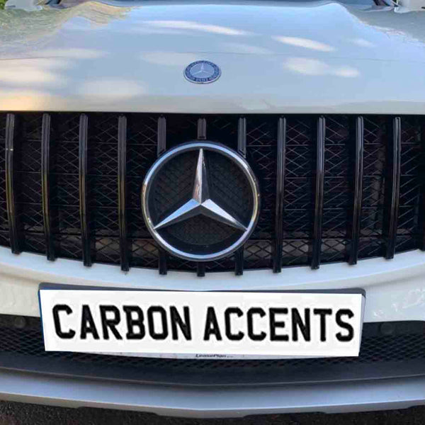 A car grille displays a prominent emblem and black vertical slats, situated on a white vehicle. The license plate reads "CARBON ACCENTS," with greenery faintly visible in the background.