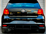 A black Volkswagen GTI is parked, displaying its rear with dual exhausts. Red lights are illuminated, positioned in front of a metallic roller door on a paved surface.