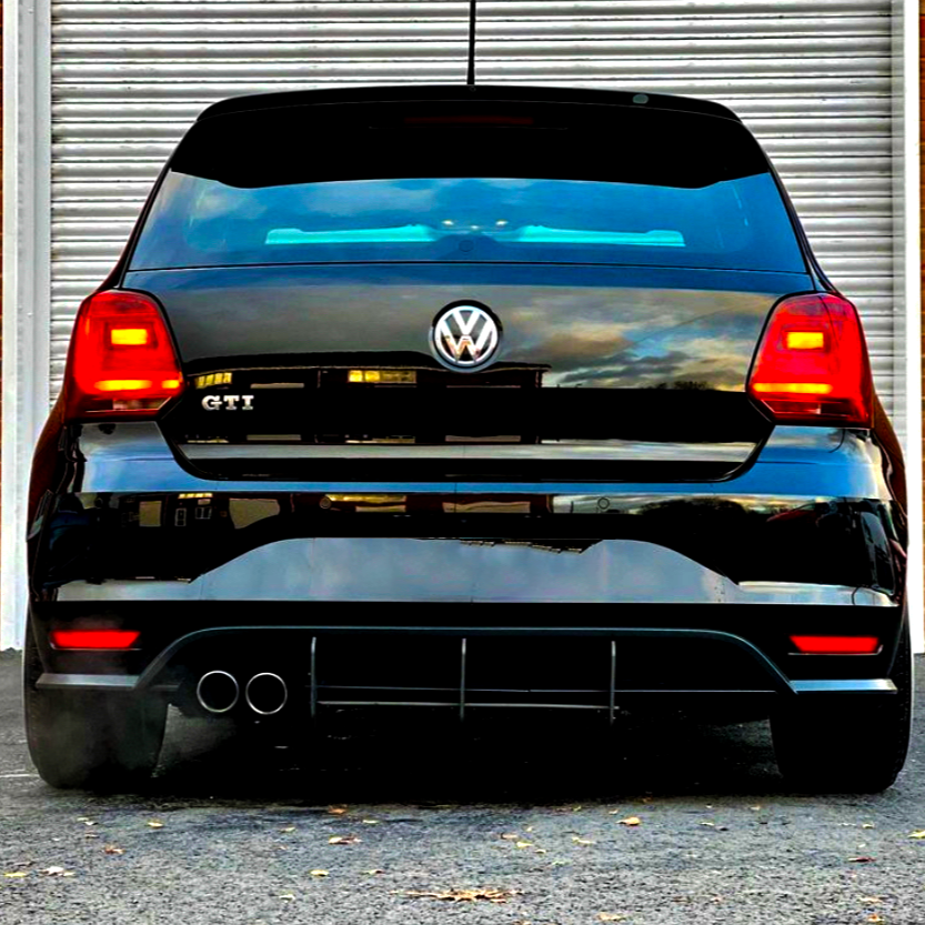 A black Volkswagen GTI is parked, displaying its rear with dual exhausts. Red lights are illuminated, positioned in front of a metallic roller door on a paved surface.