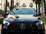A black Mercedes-Benz car with illuminated headlights is parked on a paved surface. Tall palm trees are in the background under a cloudy sky.
