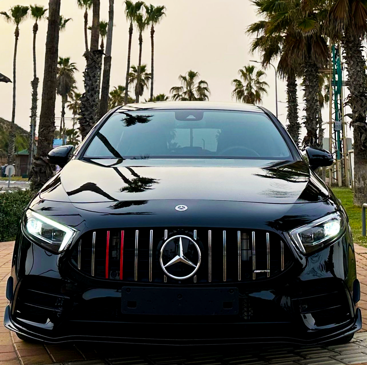 A black Mercedes-Benz car with illuminated headlights is parked on a paved surface. Tall palm trees are in the background under a cloudy sky.