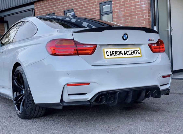 A white BMW car with black rims is parked outside a brick building. The rear license plate reads "CARBON ACCENTS."