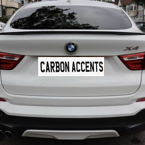 A white BMW X4 is parked on a residential street. The license plate reads "CARBON ACCENTS." Surrounding are houses and parked cars.
