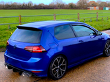 A blue car is parked, emitting exhaust, beside a grassy field with a soccer goal and wooden fencing.