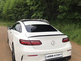 A white Mercedes-Benz coupe is parked on a dirt road, surrounded by lush green trees. The license plate reads 