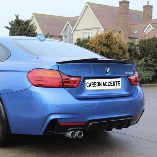 Blue sports car parked on a pavement, displaying dual exhausts and black rims. Background features a row of shrubs and residential houses. License plate reads "CARBON ACCENTS."