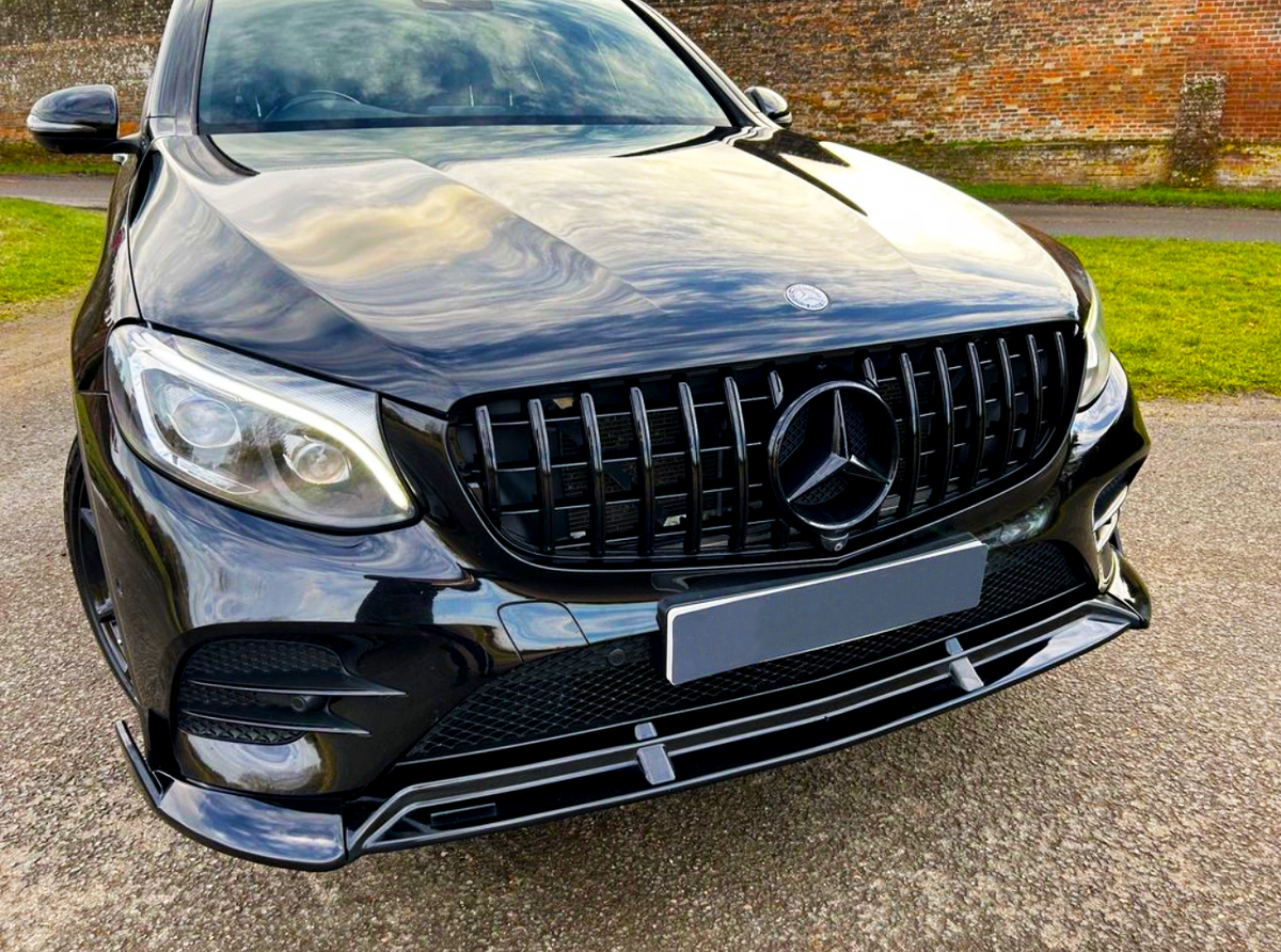 A black SUV with a prominent front grille featuring a three-pointed star emblem is parked on a paved surface, surrounded by grass and a brick wall in the background.