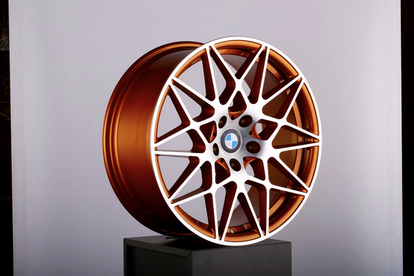 A bronze alloy wheel with intricate white spokes rests upright on a black pedestal, set against a plain white backdrop. In the center, a circular logo is visible.