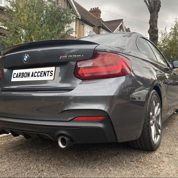 A gray BMW M235i parked on a residential street, showcasing dual exhausts and a rear spoiler. The license plate reads "CARBON ACCENTS." Nearby, brick houses and trees line the street.