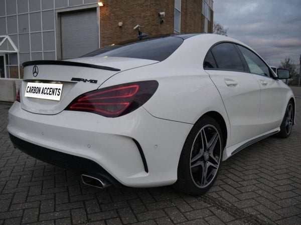A white Mercedes-Benz AMG sedan is parked on a brick-paved area beside a building. The license plate reads "CARBON ACCENTS." The environment is overcast.