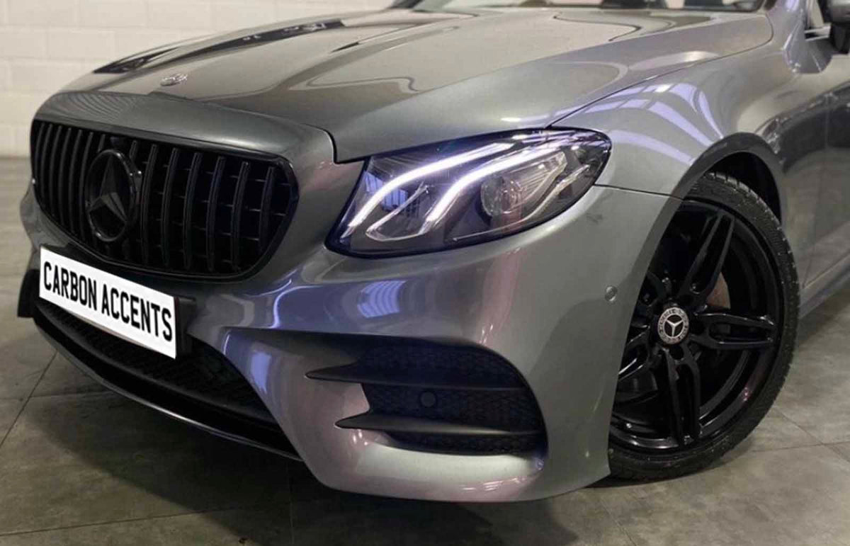 A silver car with illuminated headlights is parked indoors. A prominent grille features a star emblem. The license plate reads "CARBON ACCENTS." Black alloy wheels enhance the sleek design.