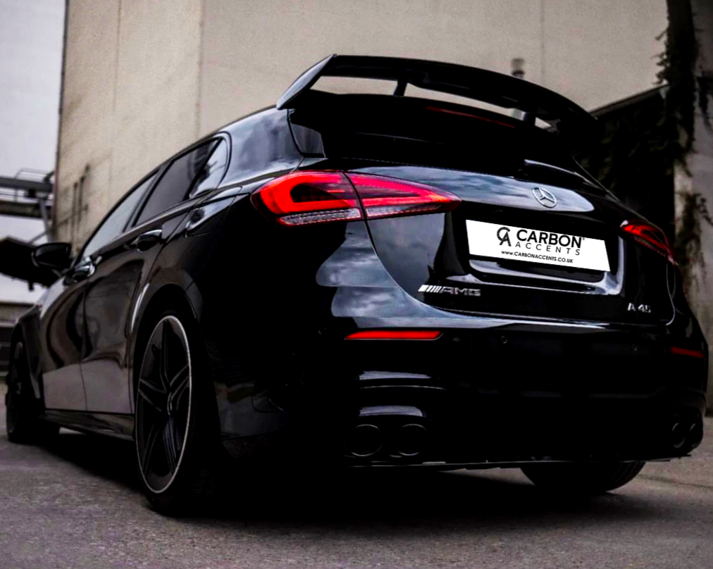A black Mercedes AMG car is parked on a street, showcasing its rear and distinctive spoiler. The license plate reads "CARBON ACCENTS," surrounded by urban buildings.