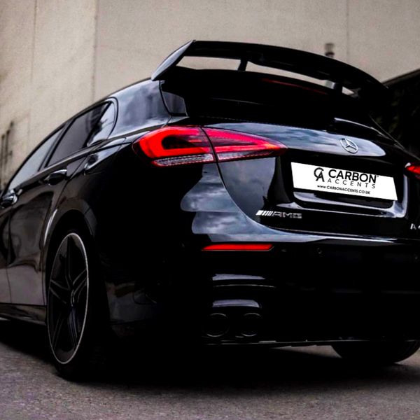 A black Mercedes AMG car is parked on a street, showcasing its rear and distinctive spoiler. The license plate reads "CARBON ACCENTS," surrounded by urban buildings.
