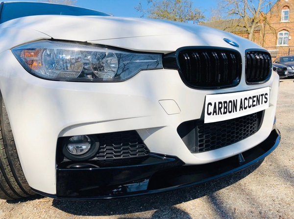 A white car with sleek black grille and headlight details sits in a sunlit parking area near trees and a brick building. The license plate reads "CARBON ACCENTS".