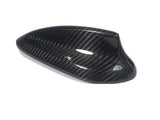 A carbon fiber car mirror cover, featuring a glossy, black textured surface, rests against a plain white background, highlighting its sleek design and curved aerodynamic shape.