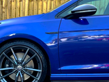 A blue car with a shiny Volkswagen logo on the wheel stands still, next to a wooden fence, reflecting light on its glossy surface.